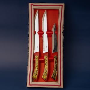 Può includere: Un set di quattro coltelli da bistecca in una scatola decorativa rossa e oro. I coltelli hanno manici rossi con filigrana dorata e lame argentate. La scatola è rossa con un bordo a strisce bianche e rosse.