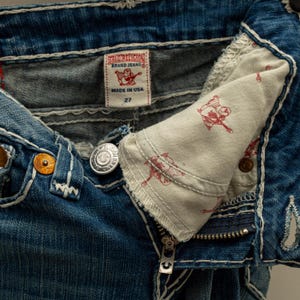 Puede incluir: Primer plano de unos vaqueros azules con costuras blancas. Los vaqueros tienen cierre de bot&oacute;n, cremallera y una etiqueta que dice "TRUE RELIGION BRAND JEANS MADE IN USA 27". El forro interior del bolsillo tiene un estampado gr&aacute;fico rojo.