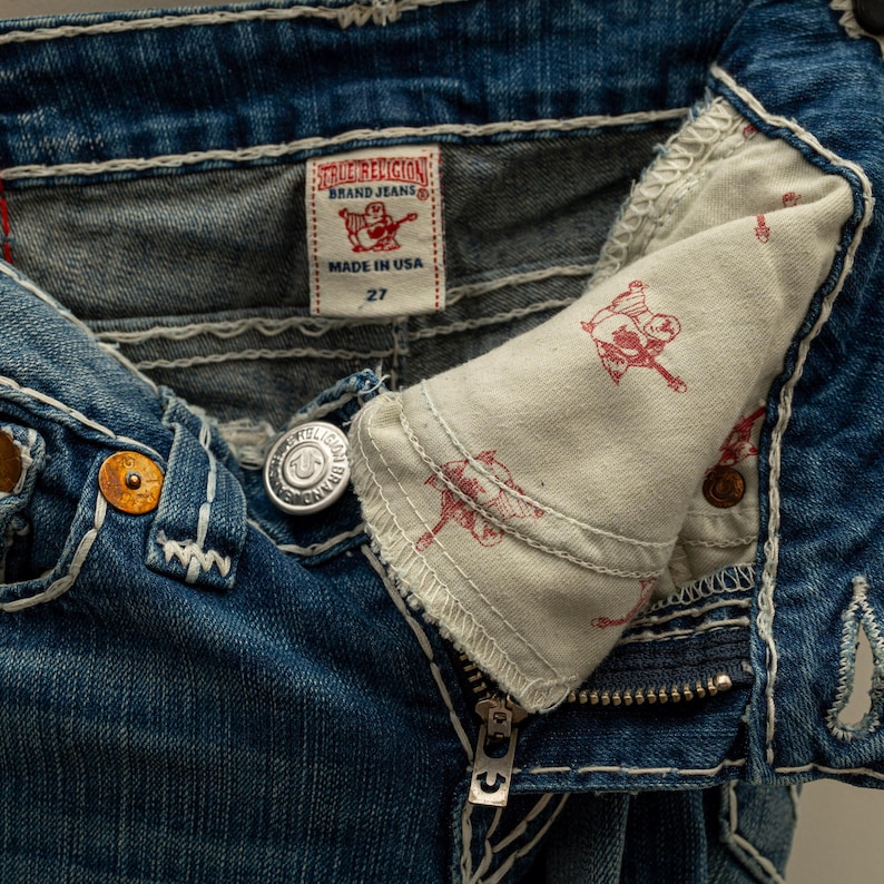Puede incluir: Primer plano de unos vaqueros azules con costuras blancas. Los vaqueros tienen cierre de bot&oacute;n, cremallera y una etiqueta que dice "TRUE RELIGION BRAND JEANS MADE IN USA 27". El forro interior del bolsillo tiene un estampado gr&aacute;fico rojo.