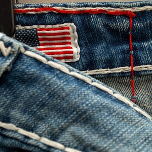 Puede incluir: Primer plano de tela vaquera azul con costuras blancas y un peque&ntilde;o parche bordado con la bandera estadounidense. La bandera presenta rayas rojas y blancas y un campo azul con estrellas blancas. Las costuras rojas acent&uacute;an la mezclilla.