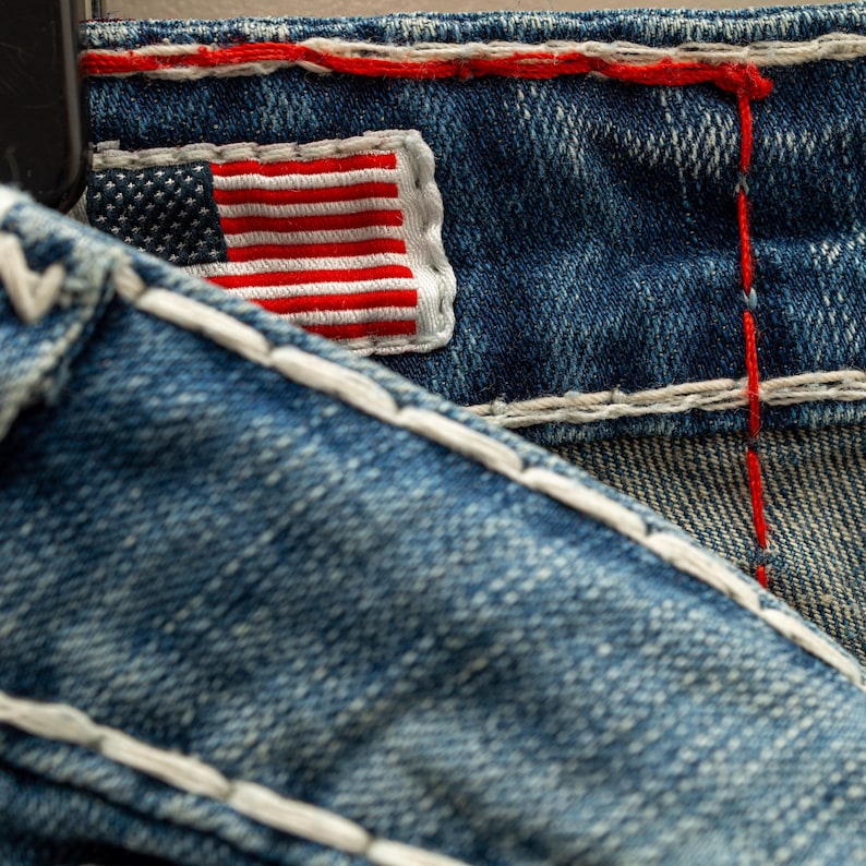 Puede incluir: Primer plano de tela vaquera azul con costuras blancas y un peque&ntilde;o parche bordado con la bandera estadounidense. La bandera presenta rayas rojas y blancas y un campo azul con estrellas blancas. Las costuras rojas acent&uacute;an la mezclilla.