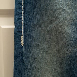 Puede incluir: Primer plano de tela vaquera azul con costuras blancas a lo largo de la costura. El denim tiene un aspecto descolorido, con &aacute;reas m&aacute;s claras que crean un aspecto desgastado. La costura es gruesa y visible.