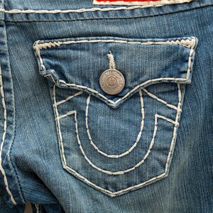 Puede incluir: Primer plano de un bolsillo de vaqueros azul claro. El bolsillo presenta un dise&ntilde;o cosido en blanco y un bot&oacute;n plateado con el texto "True Religion". Los vaqueros tienen costuras blancas.