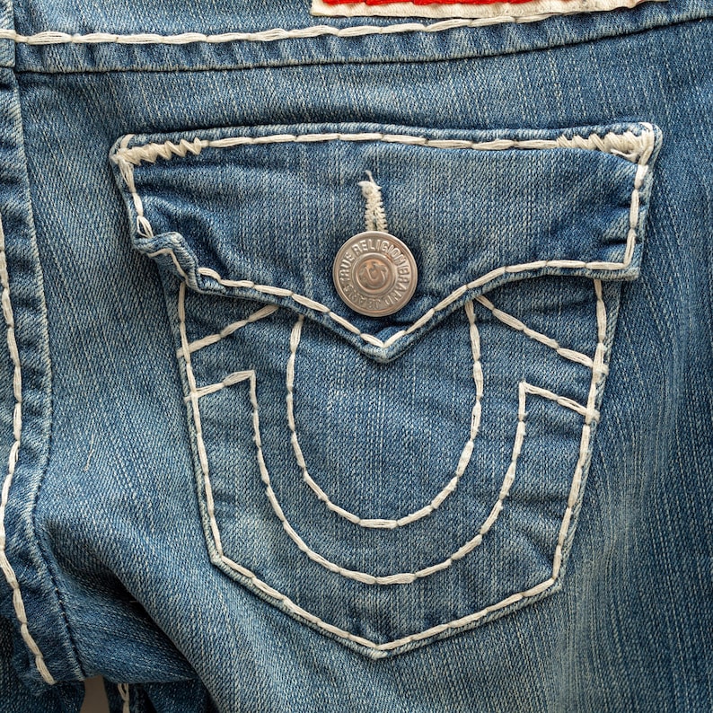 Puede incluir: Primer plano de un bolsillo de vaqueros azul claro. El bolsillo presenta un dise&ntilde;o cosido en blanco y un bot&oacute;n plateado con el texto "True Religion". Los vaqueros tienen costuras blancas.
