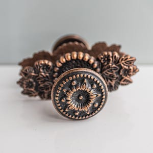 Op de afbeelding: Een koperkleurige metalen ladeknop met een ronde knop aan de voorzijde voorzien van een verhoogd bloemsterpatroon en een kralenrand. De basisplaat is langwerpig met geschulpte randen en gedetailleerde bloemmotieven in reliëf aan beide zijden.