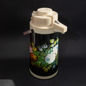 Puede incluir: Termo vintage negro y crema con diseño floral. El termo presenta una tapa y boquilla de color crema, con un cuerpo negro adornado con coloridas flores pintadas y hojas verdes. El termo tiene un asa para facilitar su transporte.
