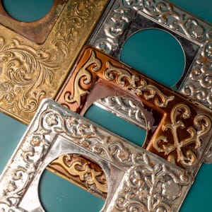 Könnte beinhalten: Nahaufnahme mehrerer verzierter Lichtschalterabdeckungen aus Metall in verschiedenen Farben. Die Abdeckungen weisen detaillierte, geprägte Blumen- und Scrollwork-Designs auf. Zu den Farben gehören Gold, Silber und Kupfer, mit einem türkisfarbenen Hintergrund.