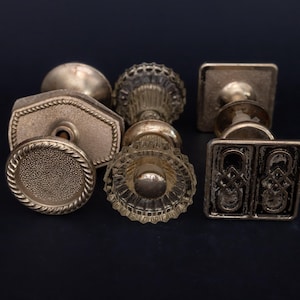 Puede incluir: Una colección de pomos de puerta antiguos de latón en varias formas, incluyendo redondos, hexagonales y cuadrados. Los pomos tienen diseños y texturas intrincados, con una apariencia cálida y envejecida. Los pomos están dispuestos sobre un fondo oscuro.