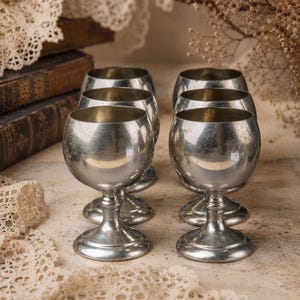 Könnte beinhalten: Ein Set aus sechs silberfarbenen Metallkelchen mit abgerundeten Kuppen, schlanken Stielen und runden Füßen. Jeder Kelch hat eine glatte, reflektierende Oberfläche und eine klassische Kelchform, angeordnet in zwei Reihen zu je drei Stück.