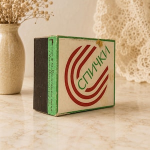 Pode incluir: Uma caixa de fósforos retangular com frente cor de creme, um gráfico circular vermelho e o texto cirílico verde "СПИЧКИ". A lateral é verde com texto preto e uma superfície de fricção castanho-escura. Contém um pequeno resíduo adesivo amarelo.