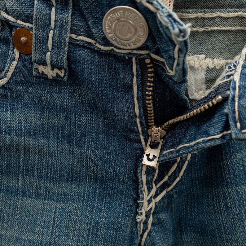 Puede incluir: Primer plano de unos vaqueros de mezclilla azules con una cremallera parcialmente abierta. Los vaqueros presentan costuras blancas, un bot&oacute;n met&aacute;lico y un tirador de cremallera plateado con un dise&ntilde;o de herradura. La tela tiene un aspecto desgastado.