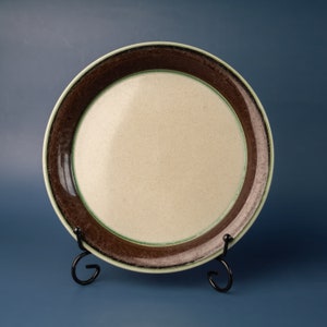 Puede incluir: Un plato redondo de cerámica con un centro color crema y un borde marrón oscuro. Una fina línea verde separa el borde marrón del centro crema. El plato se exhibe sobre un soporte de metal negro.