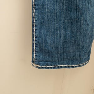 Puede incluir: Primer plano de unos vaqueros de mezclilla azules con costuras blancas a lo largo de las costuras y el dobladillo. Los vaqueros tienen un aspecto cl&aacute;sico y desgastado, con textura visible y decoloraci&oacute;n sutil. Las costuras proporcionan un detalle contrastante.