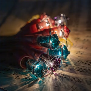 May include: A string of decorative light bulbs featuring faceted, jewel-toned glass covers in shades of teal, red, and yellow. The bulbs have a distressed, metallic finish and are arranged in a cluster, emitting a warm glow from within.