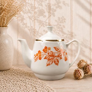 May include: A white ceramic teapot featuring a delicate orange botanical print of leaves and small flowers on its side. The teapot has a matching lid with a small knob handle, a curved spout, a side handle, and a thin gold rim around the top edge.