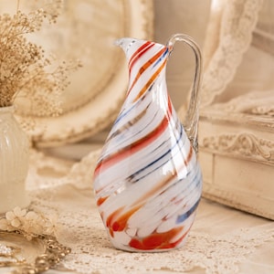 May include: A glass pitcher featuring a white base with swirling patterns of red, orange, and blue. The vessel has a narrow neck, a flared rim, and a clear glass handle attached to the side, showcasing a smooth, polished finish throughout.