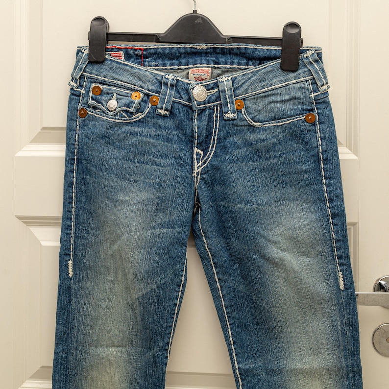 Puede incluir: Vaqueros de mezclilla azul con costuras blancas y cierre de bot&oacute;n. Los vaqueros tienen bolsillos delanteros y traseros, y est&aacute;n colgados de una percha negra. Los vaqueros tienen un lavado descolorido y est&aacute;n ligeramente desgastados.