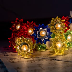 May include: A string of decorative fairy lights featuring translucent, star-shaped covers in vibrant shades of red, blue, yellow, and green. Each plastic flower-like cover houses a small, glowing light bulb, creating a warm, illuminated display.