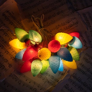 May include: A string of decorative lights featuring translucent, pinecone-shaped bulbs in vibrant shades of red, yellow, green, and blue. The bulbs are textured with a cross-hatched pattern and are illuminated, casting a warm glow from within.