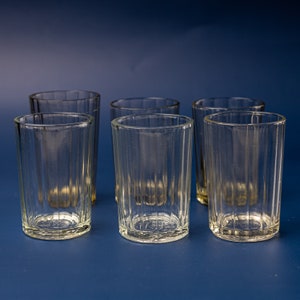 May include: Six clear, ribbed drinking glasses arranged on a blue surface. The glasses are cylindrical with a slightly flared rim. The vertical ribbing gives them a textured appearance. The glasses are empty and appear to be made of glass.