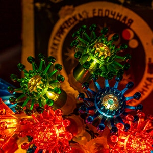 May include: A string of vintage holiday lights featuring translucent, star-shaped plastic covers in vibrant green, blue, and red. Each light cover has a spiky, multi-pointed design with small spheres at the tips, illuminated from within by warm bulbs.