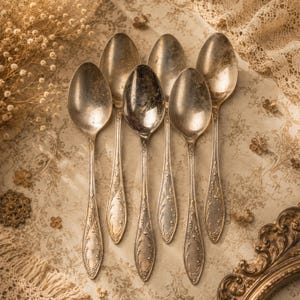 May include: A set of six silver-toned metal spoons featuring ornate, embossed floral detailing along the handles. The spoons have a tarnished, aged finish and are arranged in a fan-like pattern, showcasing their matching decorative design.