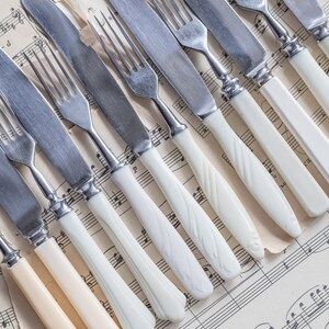 May include: An assortment of vintage silverware, including forks and knives, with white handles and stainless steel blades. The utensils are arranged diagonally on a sheet of aged musical score paper, creating a classic, elegant aesthetic.