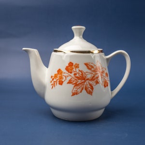 Puede incluir: Tetera de cerámica blanca con tapa, borde dorado y diseño floral naranja. La tetera tiene un pico curvo y un asa. Fondo azul liso. Ideal para servir té.