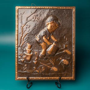 May include: A bronze-coloured relief sculpture showing a child watering plants in a garden setting. The rectangular artwork has a detailed frame and includes gardening tools, potted plants, and a tree. The theme is gardening and childhood.