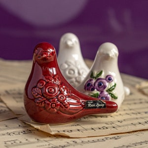 Puede incluir: Tres figuras de pájaros de cerámica. El pájaro delantero es de color rojo intenso con un diseño de rosas. Los otros dos pájaros son blancos, uno con un estampado floral y el otro con detalles de flores moradas. Los pájaros están sobre partituras, con un fondo morado.
