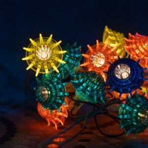Puede incluir: Una cadena de luces con forma de concha marina en colores amarillo, naranja, azul y verde. Las luces están encendidas, creando una exhibición festiva y decorativa. Las luces de cadena están sobre un fondo oscuro.