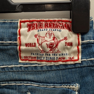 Puede incluir: Primer plano de unos vaqueros azules con costuras blancas. Una etiqueta rectangular con ribete rojo muestra el texto "TRUE RELIGION BRAND JEANS" y un dibujo de un Buda tocando la guitarra. La etiqueta tambi&eacute;n incluye las palabras "WORLD TOUR" y "SECTION BILLY SUPER T ROW SEAT."