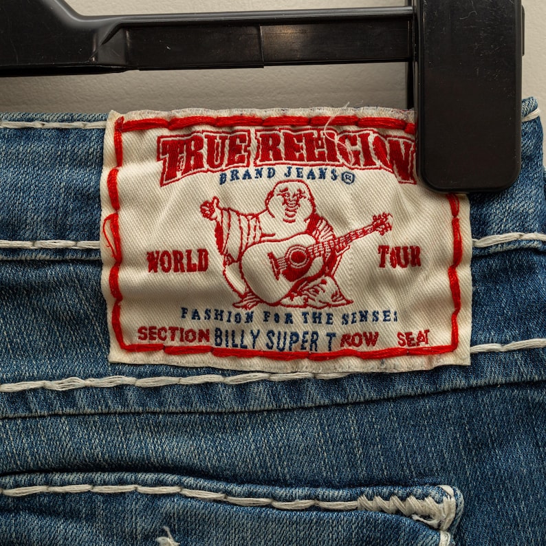 Puede incluir: Primer plano de unos vaqueros azules con costuras blancas. Una etiqueta rectangular con ribete rojo muestra el texto "TRUE RELIGION BRAND JEANS" y un dibujo de un Buda tocando la guitarra. La etiqueta tambi&eacute;n incluye las palabras "WORLD TOUR" y "SECTION BILLY SUPER T ROW SEAT."