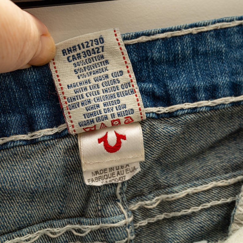 Puede incluir: Primer plano de una etiqueta de jeans de mezclilla con el texto "RN#112790 CA#30427" e instrucciones de lavado. La etiqueta tambi&eacute;n presenta un logotipo de herradura roja. Los jeans son azules con costuras blancas.