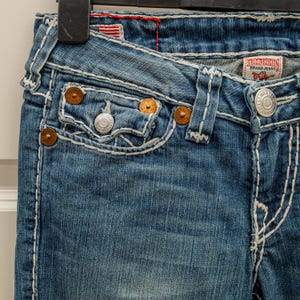 Puede incluir: Primer plano de unos vaqueros de mezclilla azules con costuras blancas. Los vaqueros presentan un dise&ntilde;o cl&aacute;sico de cinco bolsillos con remaches de color cobre y un bot&oacute;n plateado. La etiqueta de la marca True Religion es visible.