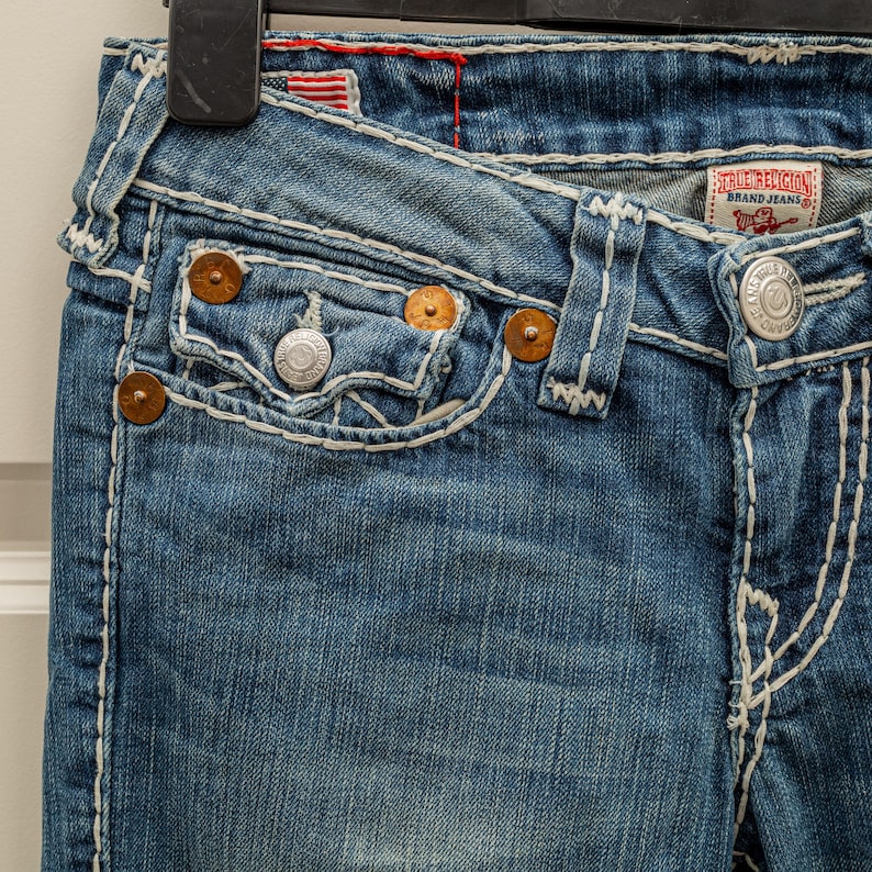 Puede incluir: Primer plano de unos vaqueros de mezclilla azules con costuras blancas. Los vaqueros presentan un dise&ntilde;o cl&aacute;sico de cinco bolsillos con remaches de color cobre y un bot&oacute;n plateado. La etiqueta de la marca True Religion es visible.