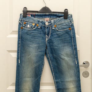 Puede incluir: Vaqueros azules de mezclilla con costuras blancas y remaches color cobre. Los vaqueros cuelgan de una percha negra contra una puerta blanca. Los vaqueros tienen un lavado descolorido y un dise&ntilde;o cl&aacute;sico de cinco bolsillos.