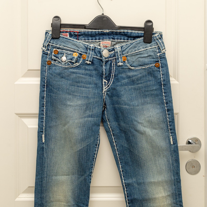 Puede incluir: Vaqueros azules de mezclilla con costuras blancas y remaches color cobre. Los vaqueros cuelgan de una percha negra contra una puerta blanca. Los vaqueros tienen un lavado descolorido y un dise&ntilde;o cl&aacute;sico de cinco bolsillos.