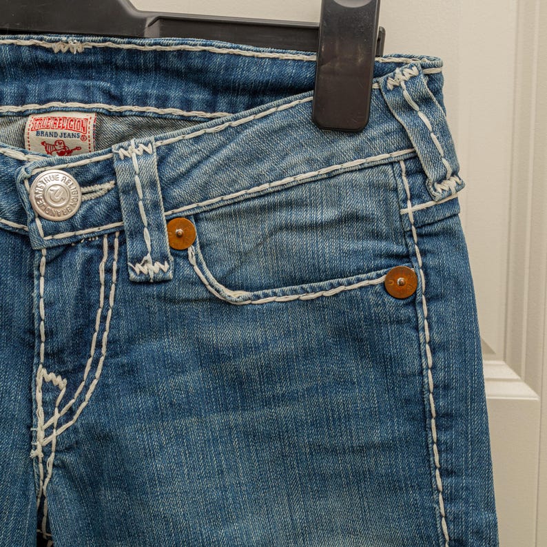 Puede incluir: Primer plano de unos vaqueros de mezclilla azules con costuras blancas y botones marrones. Los vaqueros tienen un peque&ntilde;o bolsillo y una etiqueta True Religion Brand Jeans. Los vaqueros est&aacute;n colgados de una percha negra.