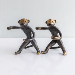 May include: Two monkey figurines crafted from dark, textured metal with polished gold-tone accents on the faces, hands, and feet. Each monkey is depicted in a dynamic, reaching pose with one arm extended forward and legs spread apart in a stance.