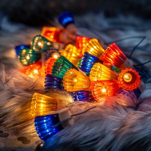 May include: A string of decorative festive lights featuring translucent, fluted, bell-shaped covers in vibrant shades of red, blue, green, and yellow. Each individual cover houses a small, glowing bulb, creating a warm, multi-coloured illumination.