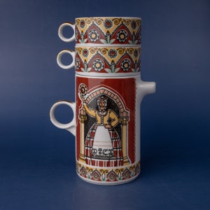 Puede incluir: Una pila de tres tazas pequeñas de cerámica blanca con bordes y asas doradas, encima de una jarra de cerámica a juego. La jarra presenta un panel rojo con una mujer con traje tradicional y la palabra "RIGA". Las tazas y la jarra tienen un diseño floral.