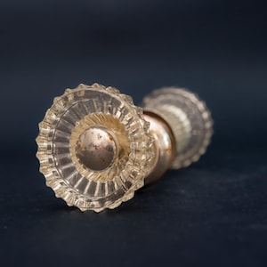 Peut inclure: Un bouton de porte en verre transparent avec un motif circulaire strié. Le bouton a une base en métal doré terni et un motif décoratif en forme d'éclat d'étoile sur le verre. Le fond est bleu foncé.