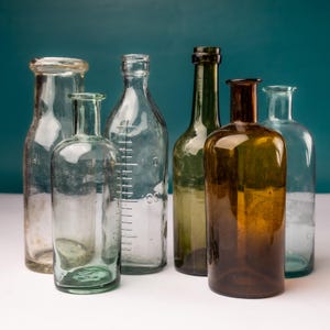 Puede incluir: Una colección de seis botellas de vidrio antiguas de diversas formas y tamaños. Las botellas son transparentes, verdes y ámbar, algunas con marcas de medición. Están dispuestas sobre una superficie blanca contra un fondo turquesa.
