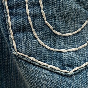 Puede incluir: Primer plano de un bolsillo de mezclilla con costuras blancas. El bolsillo es de color azul medio y las costuras son un dise&ntilde;o grueso y decorativo. La mezclilla tiene una apariencia texturizada.