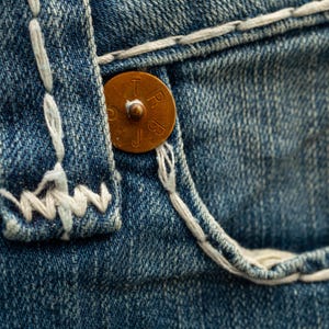 Puede incluir: Primer plano de vaqueros de mezclilla azul con un bot&oacute;n de color cobre y costuras blancas. El bot&oacute;n tiene letras en relieve. Las costuras son visibles a lo largo del bolsillo y las costuras, resaltando la textura de la tela.