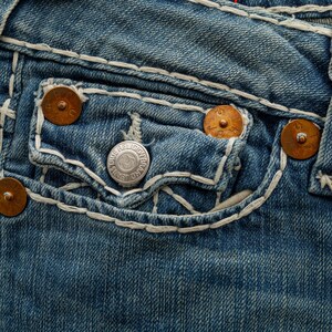 Puede incluir: Primer plano de unos vaqueros de mezclilla azul con costuras blancas y remaches color cobre. Los vaqueros tienen un bolsillo con un bot&oacute;n plateado que dice "TRUE RELIGION BRAND JEANS".