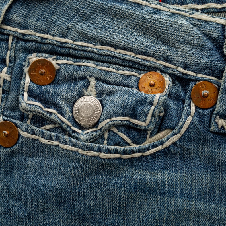 Puede incluir: Primer plano de unos vaqueros de mezclilla azul con costuras blancas y remaches color cobre. Los vaqueros tienen un bolsillo con un bot&oacute;n plateado que dice "TRUE RELIGION BRAND JEANS".