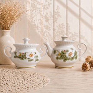 Może przedstawiać: Biały ceramiczny zestaw składający się z cukiernicy i czajnika, ozdobiony malowanym wzorem białych kwiatów, zielonych igieł sosnowych i małych czerwonych jagód. Obie części posiadają pokrywki, złote obrzeża podstawy oraz zakrzywione uchwyty.