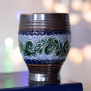 May include: A ceramic mug with a dark brown top and base, and a light blue center with green floral designs. The mug has a cylindrical shape with horizontal ridges and a glossy finish. The background is blurred with bokeh lights.